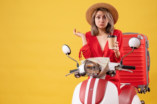 Señorita molesta en vestido rojo sosteniendo una taza de café cerca del ciclomotor