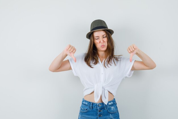 Señorita mostrando doble pulgar hacia abajo en camiseta, jeans, sombrero y mirando disgustado, vista frontal.