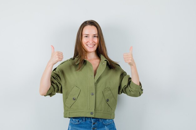 Señorita mostrando doble pulgar hacia arriba en chaqueta verde, pantalones cortos y aspecto alegre. vista frontal.