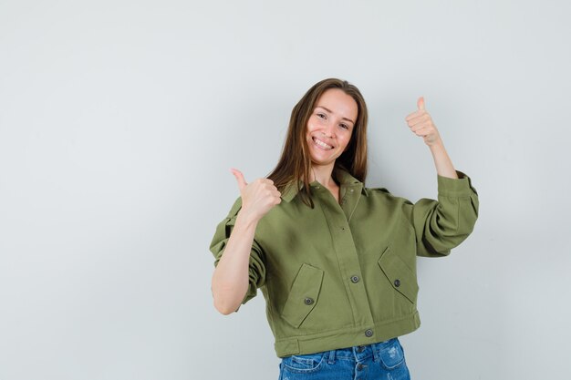 Señorita mostrando doble pulgar hacia arriba en chaqueta verde, pantalones cortos y mirando alegre. vista frontal.