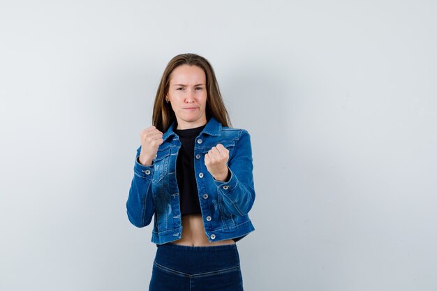 Señorita mostrando gesto de ganador en blusa, chaqueta, jeans y mirando confiado, vista frontal.