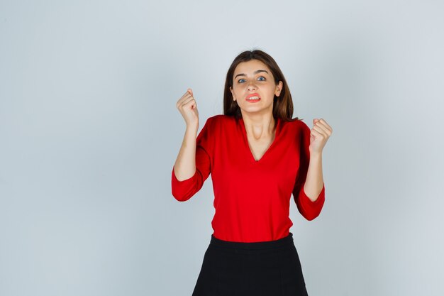 Señorita mostrando gesto ganador en blusa roja, falda y mirando dichoso