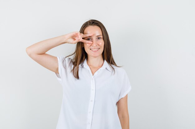 Señorita mostrando gesto de ganador en un ojo en blusa blanca y mirando feliz