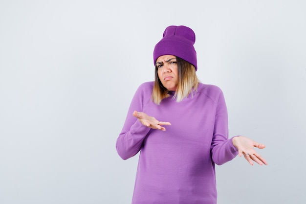 Señorita mostrando gesto de impotencia en suéter morado, gorro y mirando insatisfecho, vista frontal.