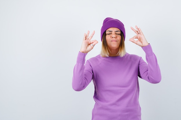 Señorita mostrando gesto de meditación en suéter morado, gorro y mirando pacífica, vista frontal.