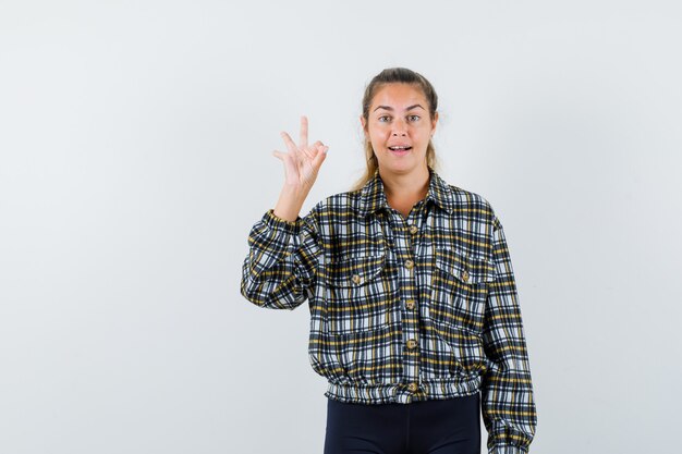 Señorita mostrando gesto ok en camisa, pantalones cortos y mirando alegre, vista frontal.