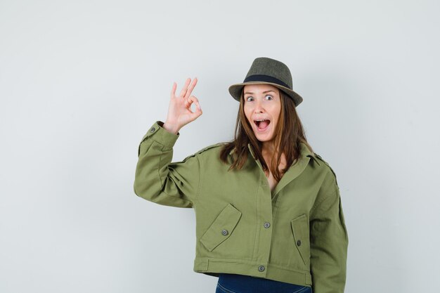 Señorita mostrando gesto ok en chaqueta pantalones sombrero y mirando feliz