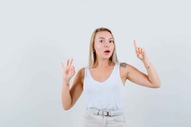 Señorita mostrando un gesto de ok mientras apunta hacia arriba en una blusa blanca y parece complacida