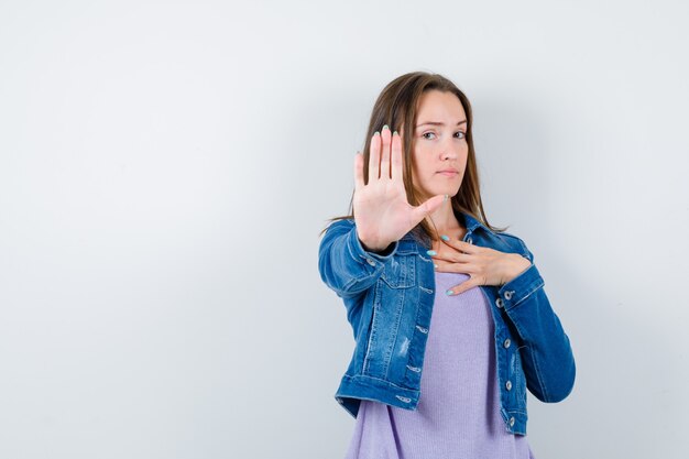 Señorita mostrando gesto de parada, manteniendo la mano en el pecho en camiseta, chaqueta y mirando serio, vista frontal.