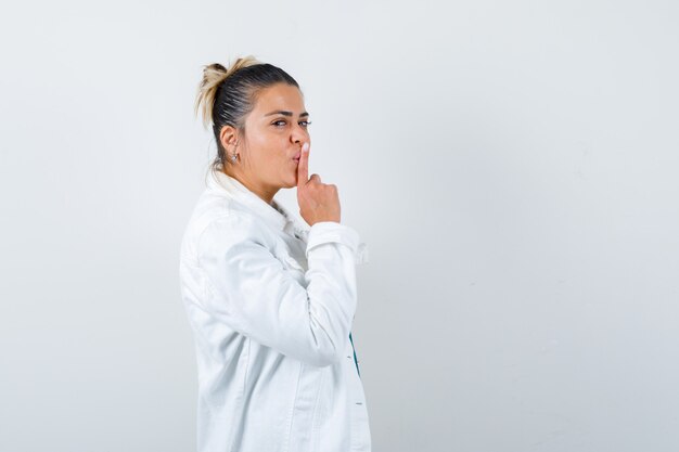 Señorita mostrando gesto de silencio en camisa, chaqueta blanca y mirando con cuidado. vista frontal.