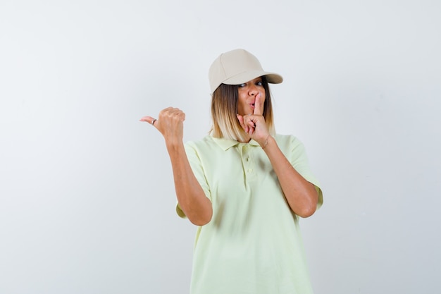 Señorita mostrando gesto de silencio mientras apunta hacia el lado izquierdo con el pulgar en la camiseta, gorra y mirando confiado, vista frontal.