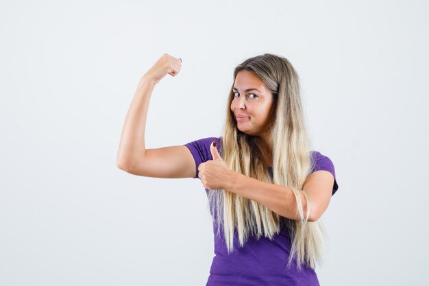 Señorita mostrando los músculos con el pulgar hacia arriba en camiseta violeta y luciendo confiada. vista frontal.