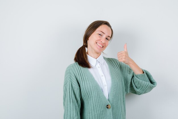 Señorita mostrando el pulgar hacia arriba en blusa, chaqueta de punto y mirando alegre, vista frontal.