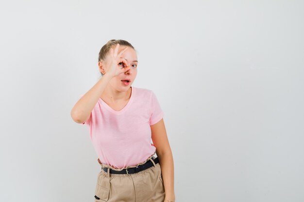 Señorita mostrando signo ok en el ojo en camiseta y pantalón y mirando curioso