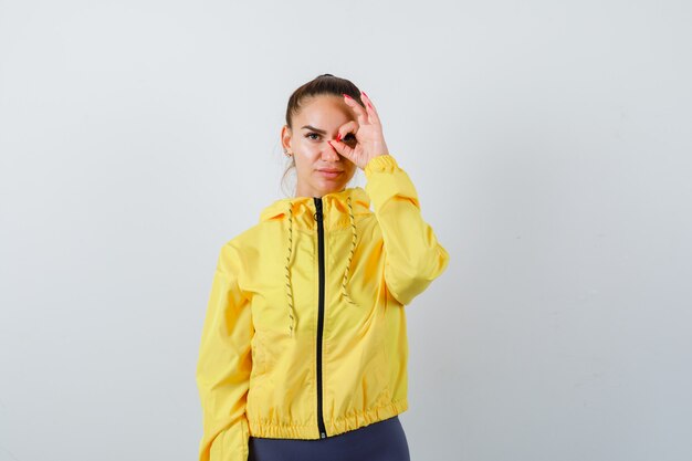 Señorita mostrando signo ok en el ojo con chaqueta amarilla y mirando confiado, vista frontal.