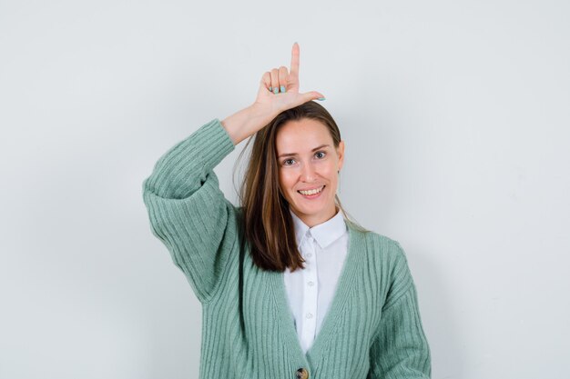 Señorita mostrando signo de perdedor en camisa, chaqueta de punto y mirando alegre. vista frontal.