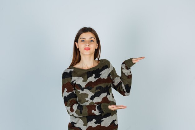 Señorita mostrando signo de tamaño en suéter, falda y mirando complacido