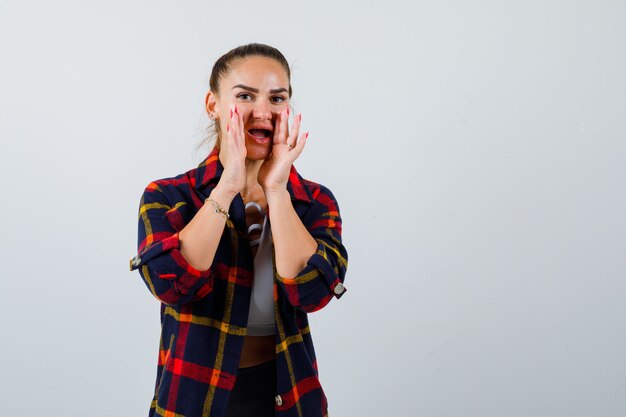 Señorita en la parte superior, camisa a cuadros con las manos cerca de la boca para decir el secreto y mirando emocionada, vista frontal.