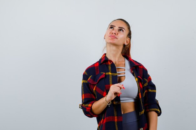 Señorita en la parte superior, camisa a cuadros posando mientras mira hacia arriba y mira pensativa, vista frontal.