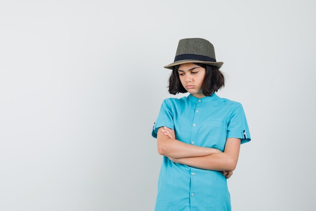 Señorita de pie con los brazos cruzados en camisa azul, sombrero y mirando ofendido