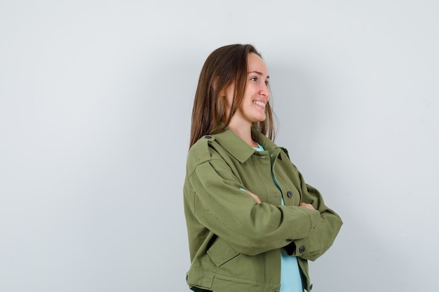 Señorita de pie con los brazos cruzados mientras mira a otro lado en camiseta, chaqueta y parece alegre. vista frontal.