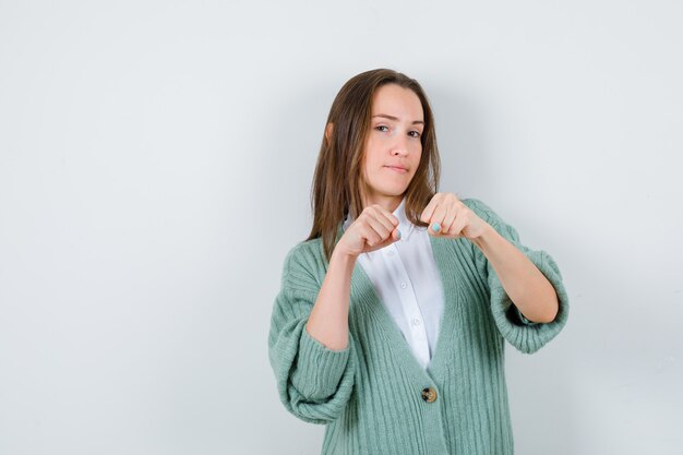 Señorita de pie en pose de lucha en camisa, chaqueta de punto y mirando confiado, vista frontal.