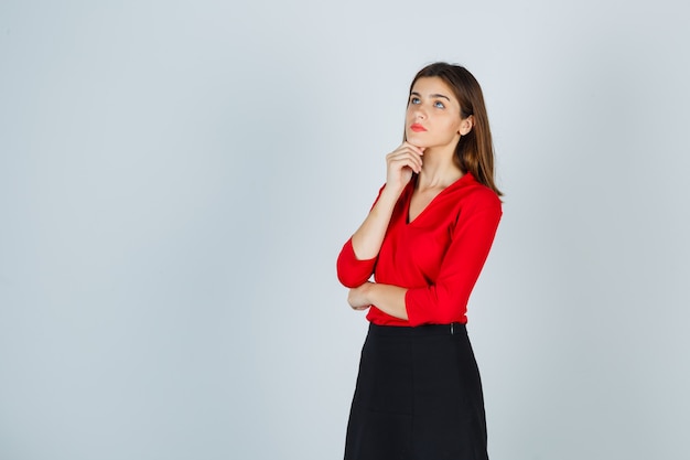 Señorita de pie en pose de pensamiento, apoyando la barbilla en el puño en blusa roja