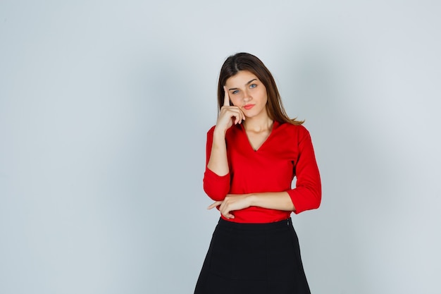 Señorita de pie en pose de pensamiento en blusa roja, falda y mirando pensativo