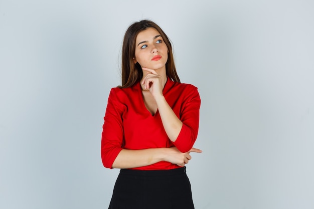 Señorita de pie en pose de pensamiento en blusa roja, falda y mirando pensativo