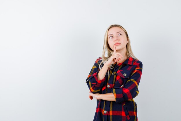 Señorita de pie en pose de pensamiento en camisa a cuadros y mirando indeciso. vista frontal.