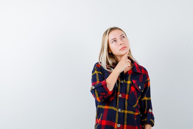 Señorita de pie en pose de pensamiento en camisa a cuadros y mirando pensativo, vista frontal.