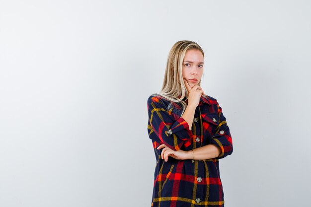 Señorita de pie en pose de pensamiento en camisa a cuadros y mirando pensativo, vista frontal.