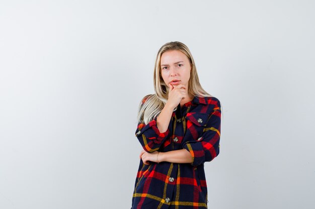 Señorita de pie en pose de pensamiento en camisa a cuadros y mirando pensativo, vista frontal.