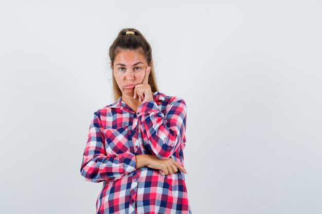 Señorita de pie en pose de pensamiento en camisa a cuadros y mirando triste. vista frontal.