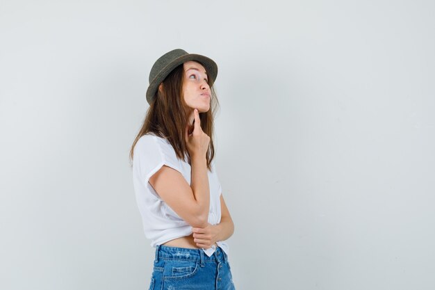 Señorita de pie en pose de pensamiento en camiseta, jeans, sombrero y mirando vacilante, vista frontal.