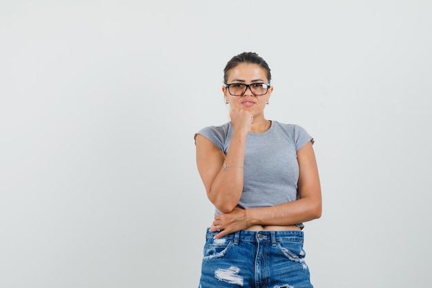 Señorita de pie en pose de pensamiento en camiseta, pantalones cortos y mirada inteligente.