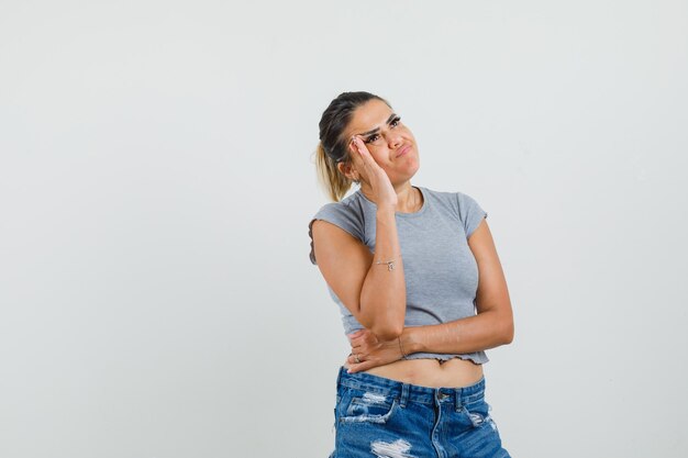 Señorita de pie en pose de pensamiento en camiseta, pantalones cortos y mirando vacilante.