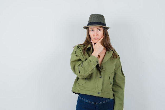 Señorita de pie en pose de pensamiento en chaqueta pantalones sombrero y mirando serio