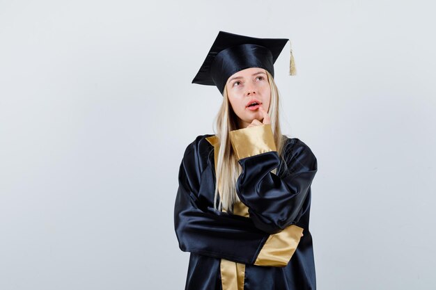 Señorita de pie en pose de pensamiento en traje académico y mirando pensativo