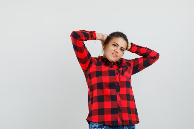 Señorita posando con las manos detrás de la cabeza en camisa a cuadros, pantalones cortos y aspecto encantador.