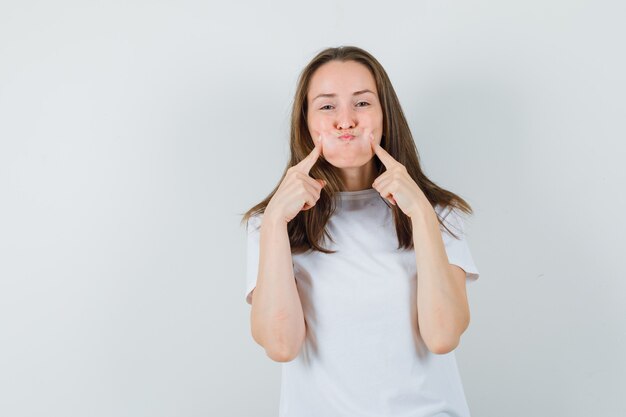 Señorita presionando los dedos en las mejillas sopladas en camiseta blanca