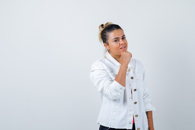 Señorita con puño en la barbilla en camiseta, chaqueta blanca y mirando confiado, vista frontal.