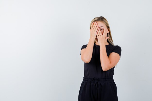 Señorita que cubre los ojos con la mano mientras mira a través de los dedos en camiseta, pantalones y parece asustada. vista frontal.