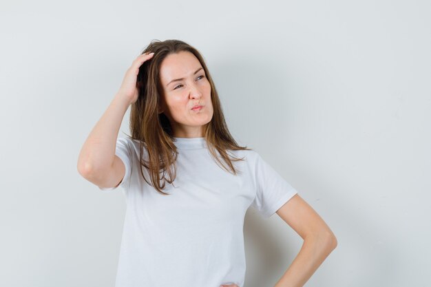 Señorita rascándose la cabeza en camiseta blanca y mirando vacilante