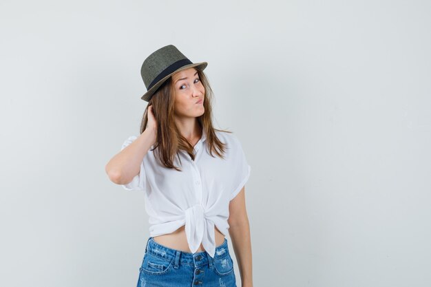 Señorita rascándose la cabeza en camiseta, jeans, sombrero y mirando pensativo, vista frontal.