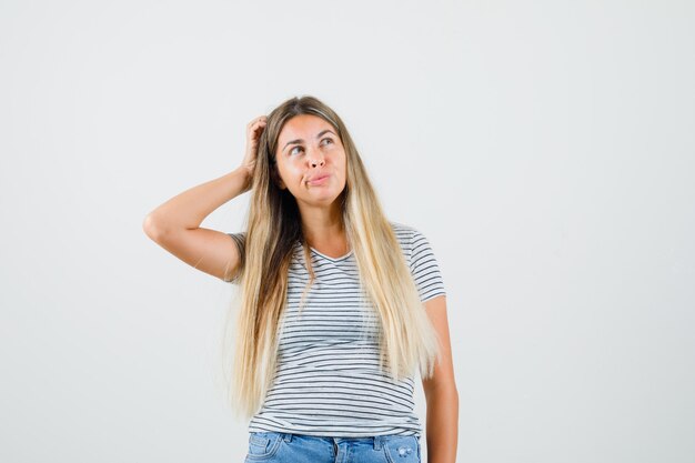 Señorita rascándose la cabeza mientras mira a otro lado en camiseta y mira pensativa, vista frontal.