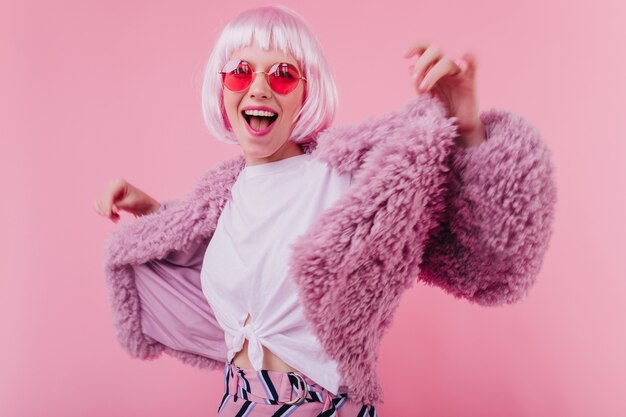Señorita refinada en corto periwig bailando en la pared rosa. Riendo a adorable niña con gafas de sol posando en elegante chaqueta de piel