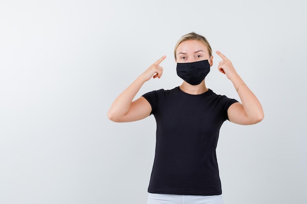 Señorita señalando la cabeza en camiseta, pantalón, máscara médica y luciendo orgullosa