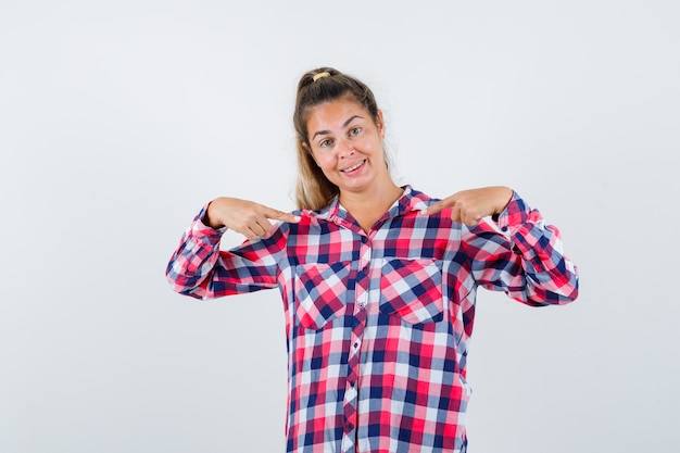 Señorita señalando a sí misma en camisa a cuadros y luciendo orgullosa. vista frontal.