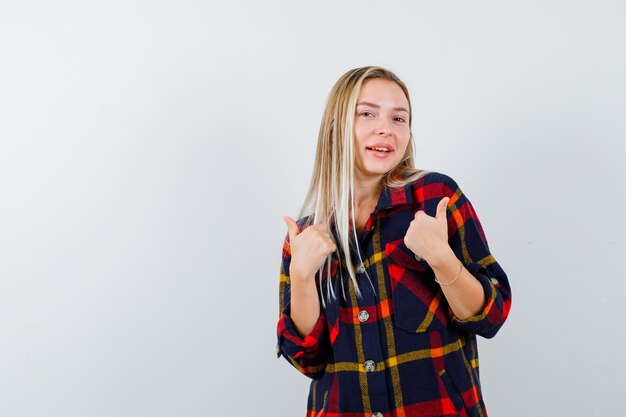 Señorita señalando a sí misma en camisa a cuadros y luciendo orgullosa. vista frontal.